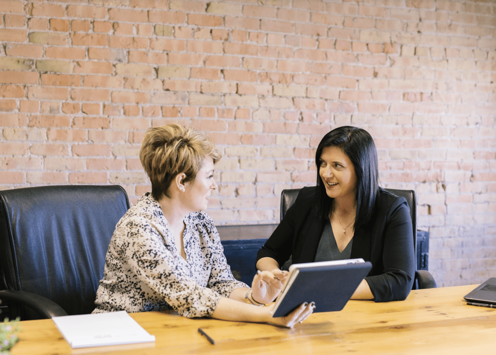 Two smartly dressed women are sat at a desk having a meeting whilst using a tablet.