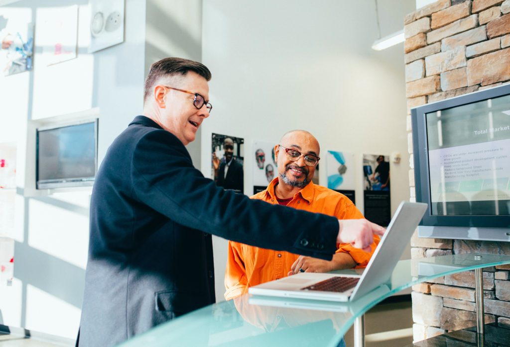 Two employees working from a laptop. One employee is standing and pointing to a laptop screen whilst the other is sat down and smiling whilst looking at the screen.